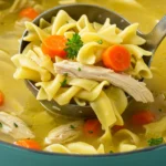 Ladle lifting chicken noodle soup with egg noodles, carrots, and parsley from a teal-blue pot