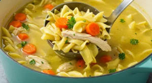 Ladle lifting chicken noodle soup with egg noodles, carrots, and parsley from a teal-blue pot