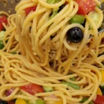 Overhead angled view of pasta salad with spaghetti, red onion, bell peppers, cucumber, tomatoes, and olives, with serving utensil in action