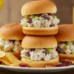 Three cranberry chicken salad sliders on a white plate with potato chips, garnished with green onions, nuts, and cranberries.