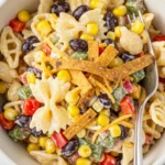 Bright overhead image of a small round white bowl with Mexican pasta salad, including corn, black beans, red bell peppers, shredded cheese, crispy tortilla strips, and a silver fork on marble