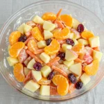 Top-down view of a glass bowl filled with vibrant carrot apple salad featuring mandarin oranges and cranberries on a striped placemat