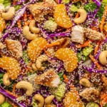 Overhead shot of a white bowl filled with Chinese chicken salad, including shredded lettuce, purple cabbage, carrots, mandarin oranges, cashews, roasted chicken, and sesame seeds, on a mottled gray-blue surface.