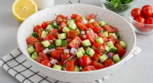 Overhead view of a vibrant cucumber tomato salad with red onion and dill in a white bowl, surrounded by fresh ingredients on a checkered cloth.