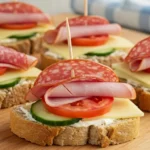 Close-up of open-faced tea club sandwiches topped with ham, salami, cheese, tomato, and cucumber on a wooden board