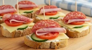 Close-up of open-faced tea club sandwiches topped with ham, salami, cheese, tomato, and cucumber on a wooden board