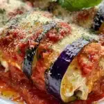 Eggplant rollatini with melted cheese and marinara sauce on a white plate, garnished with fresh basil