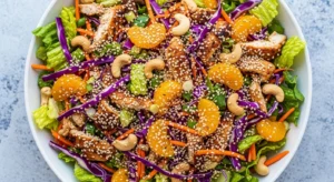 Close-up of a fresh Chinese chicken salad in a white bowl, featuring shredded lettuce, purple cabbage, carrots, cashews, mandarin oranges, grilled chicken, and sesame seeds on a light gray-blue surface.