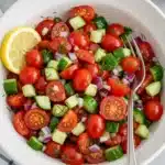 Cucumber, cherry tomatoes, and red onion mixed with dill in a speckled white bowl on a gray surface with a checkered tablecloth and fresh garnishes.