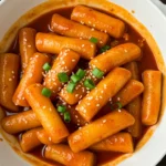 White bowl of Korean spicy tteokbokki topped with sesame seeds and chopped scallions, placed on dark wood with a striped napkin