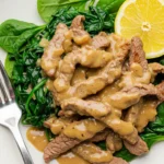 Plate of beef stir-fry with spinach in light brown creamy sauce, lemon wedge on top, on marble countertop