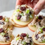 Hand lifting a thin apple slice topped with creamy cranberry chicken salad mixed with celery and pecans from a white plate