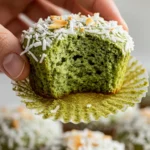 A woman's hand gently lifting a matcha coconut muffin topped with shredded coconut and toasted flakes, showcasing its pale green crumb.