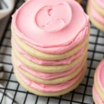 Close-up of a crumbl cookie with creamy pink frosting stacked on a cooling rack beside a striped towel.