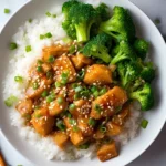 Glazed teriyaki chicken slices on a bed of white rice, topped with chopped green onions and sesame seeds, paired with fresh steamed broccoli on a white plate.