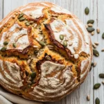 Artisan round oat bread with golden cracked crust and embedded sunflower and pumpkin seeds on a rustic wooden surface