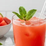 A clear round glass of watermelon juice with ice and mint on a marble countertop, lime wedges and cubed watermelon beside it