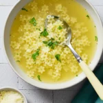 Star-shaped pasta in a transparent golden broth, served in an off-white bowl with Parmesan cheese and parsley garnish, a spoon resting in the soup.