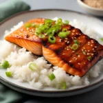 Glazed teriyaki salmon fillet on white rice, topped with sesame seeds and green onions on a white plate with dark rim